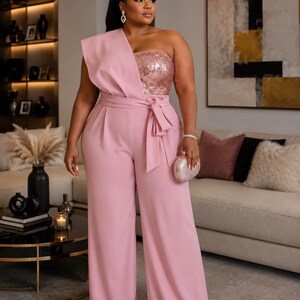 May include: A light pink one-shoulder jumpsuit featuring a sequined bodice, a wide-leg silhouette, and a matching fabric tie-waist belt. The garment includes a structured drape over one shoulder and is paired with a small, round, metallic clutch bag.