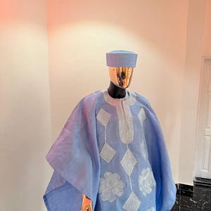 以下が含まれることがあります： 金色の頭と手を持つマネキンに展示された、水色の伝統的な衣装。チュニックと帽子が含まれています。チュニックには、白いダイヤモンドと花の刺繍模様があります。この衣装は軽量の生地で作られています。