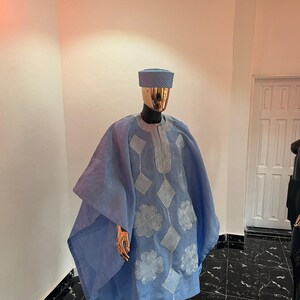 以下が含まれることがあります： マネキンに展示された、水色の伝統的な衣装。刺繍された花柄とひし形模様のチュニック、お揃いのパンツ、帽子が含まれています。この衣装は軽量の生地で作られており、マネキンは金色の頭と手を持っています。
