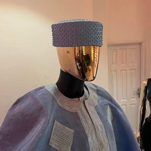 以下が含まれることがあります： 金色の頭のマネキンに展示された、水色の伝統的な衣装。チュニックとマッチする帽子が含まれています。チュニックは襟と前面に銀色の刺繍が施され、ひし形のパッチが付いています。