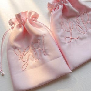 May include: Two light pink drawstring pouches made of a smooth fabric. Each bag features a matching pink embroidered monogram on the front. The left pouch displays the letters GH, while the right pouch displays the letters LB in a cursive script.