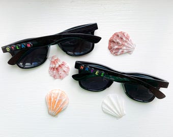 May include: Two pairs of black sunglasses with colorful beaded names, "DYLAN" and "HAYES", displayed on the frames. The sunglasses are surrounded by various seashells in shades of pink, orange, and white, set against a white background.