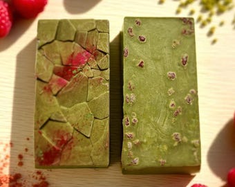 May include: Two rectangular green bars with red accents, possibly soap or chocolate, are displayed on a light wooden surface. Fresh red raspberries and a scattering of red powder and green granules surround the bars, suggesting a natural ingredient theme.