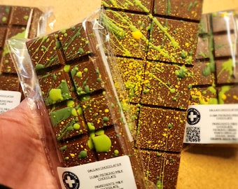 Puede incluir: Se muestran varias barras de chocolate con leche y pistachos de Dubái. El chocolate marrón oscuro está decorado con salpicaduras verdes y amarillas. Cada barra está empaquetada en plástico transparente, con una etiqueta que incluye la marca y los ingredientes.