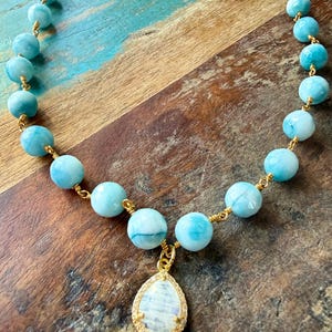 May include: A necklace featuring light blue, round beads strung on gold-colored wire. A teardrop-shaped pendant with a white and blue stone and gold-colored trim hangs from the center. The necklace is displayed on a wooden surface.