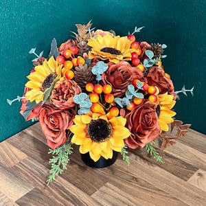 May include: A floral arrangement featuring yellow sunflowers, rust-coloured roses, brown pinecones, and bright orange berries. The bouquet includes sprigs of sage green eucalyptus and fern-like foliage, all contained within a black circular base.