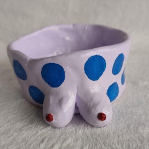 May include: A handmade ceramic bowl in a light purple hue, adorned with large blue polka dots. The bowl features two rounded protrusions with red accents. The design is whimsical and artistic, suitable for decorative use.
