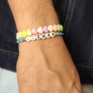 Op de afbeelding: Twee kralenarmbanden die samen worden gedragen. De bovenste armband heeft pastelkleurige hartvormige kralen in geel, roze, paars en oranje. De onderste armband bestaat uit kleine blauwe kralen en witte ronde kralen die het woord ICEMAN spellen.