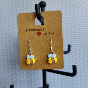 Könnte beinhalten: Handgemachte Bienenohrringe mit gelben Körpern, braunen Streifen und hellblauen Flügeln. Die Ohrringe werden auf einer braunen Karte mit dem Text "Handmade with love" präsentiert.