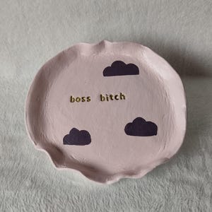 Puede incluir: Un plato de cerámica rosa claro hecho a mano con un borde festoneado. El plato presenta tres diseños de nubes moradas oscuras y el texto dorado "boss bitch". Este plato decorativo es perfecto para guardar joyas o objetos pequeños.