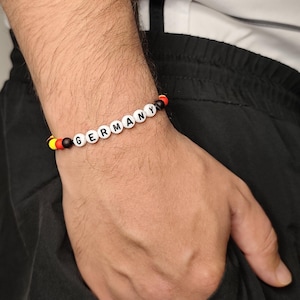 Puede incluir: Una pulsera de cuentas con cuentas esféricas blancas con el texto negro GERMANY impreso, flanqueadas por cuentas más pequeñas con los colores de la bandera alemana: negro, rojo y amarillo, dispuestas en una secuencia sobre un cordón fino.
