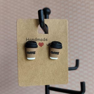 Könnte beinhalten: Handgemachte Kaffeetassen-Ohrringe. Jeder Ohrring ist eine Miniatur-Kaffeetasse in Braun mit schwarzem Deckel und dem Wort "COFFEE" auf der Seite. Die Ohrringe werden auf einer braunen Karte mit den Worten "Handmade with" und einem roten Herz präsentiert.