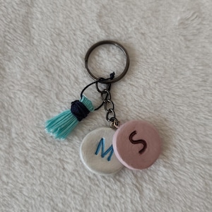 May include: A handmade keychain featuring a metal ring, chain, and two circular charms. One charm is white with a blue "M", the other is pink with a brown "S". A blue tassel adds a decorative touch.