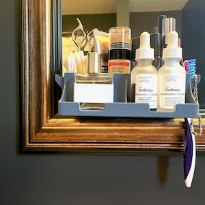 May include: A gray shelf attached to a mirror holds various beauty products. Items include bottles of The Ordinary skincare, Revlon makeup, perfume, and a toothbrush. The mirror has a decorative gold frame and is mounted on a dark gray wall.