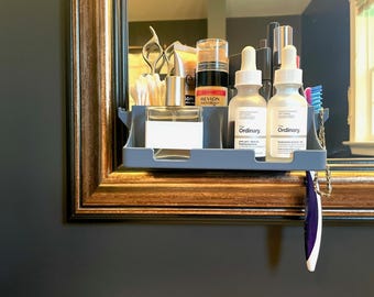 May include: A gray shelf attached to a mirror holds various beauty products. Items include bottles of The Ordinary skincare, Revlon makeup, perfume, and a toothbrush. The mirror has a decorative gold frame and is mounted on a dark gray wall.