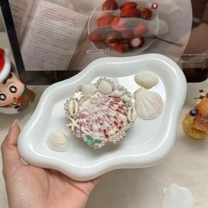 May include: A white, wavy-edged ceramic tray holds a seashell arrangement. The arrangement features a large, red and white striped shell surrounded by smaller shells, pearls, and a starfish. Additional seashells are scattered on the tray.