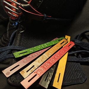 May include: A collection of colorful leather straps with embossed floral and geometric patterns. The straps, in shades of green, pink, orange, red, black, and yellow, are arranged near a black object with blue laces, likely part of martial arts gear.