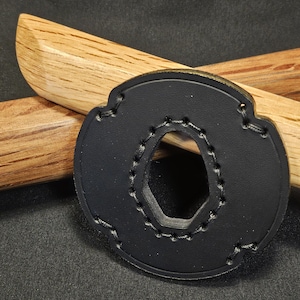 May include: A black leather tsuba, a sword guard, with a decorative shape and a central opening. The tsuba is stitched around the edges and rests in front of two wooden handles.