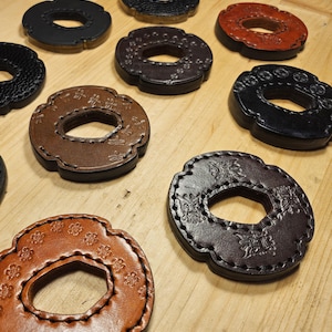 Könnte beinhalten: Eine Sammlung von Leder-Tsubas, oder Schwertschützern, in verschiedenen Braun-, Schwarz- und Rottönen. Jeder Schutz weist aufwendige geprägte Designs und dekorative Nähte an den Rändern auf. Die Schützer sind auf einer hellen Holzoberfläche angeordnet.