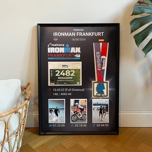 Può includere: Esposizione nera incorniciata con testo "IRONMAN FRANKFURT", dettagli della gara e medaglia del finisher. Include pettorale con "2482 BENJAMIN" e timestamp. La cornice è esposta su un pavimento in legno.