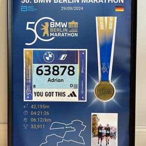 May include: Framed commemorative display of the 50th BMW Berlin Marathon, featuring a race bib, medal, and a photo of runners. The bib displays the runner's number, name, and the text "YOU GOT THIS". The medal has a gold finish.