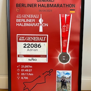 May include: Framed display featuring a red background with the text "GENERALI BERLINER HALBMARATHON" and a race bib. Includes a medal with a red ribbon, a photo, and race details. The frame is black.