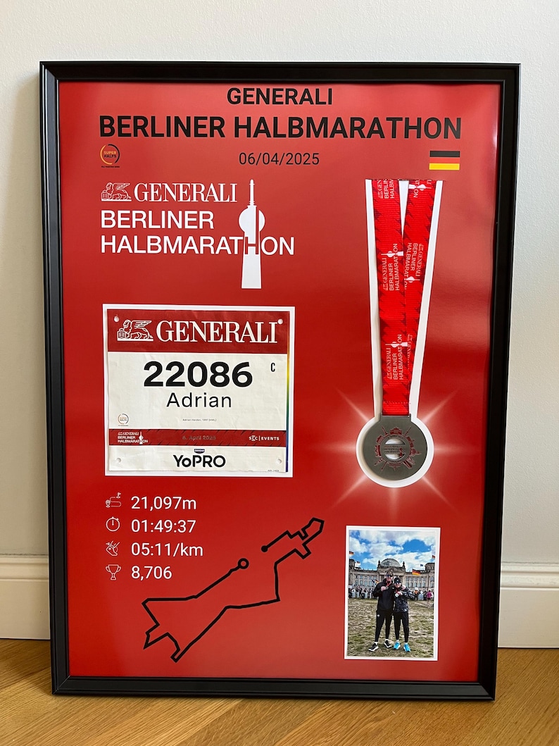 May include: Framed display featuring a red background with the text "GENERALI BERLINER HALBMARATHON" and a race bib. Includes a medal with a red ribbon, a photo, and race details. The frame is black.