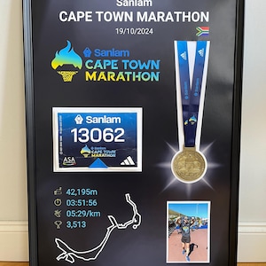 May include: Framed black display featuring a Cape Town Marathon medal, race bib, and runner's photo. The display includes the race name, date, and runner's stats: 42,195m, 03:51:56, 05:29/km, and 3,513.