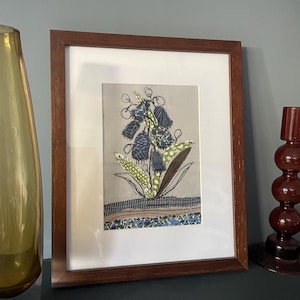 May include: Framed artwork with a floral design in blue, green, and brown. The artwork is set against a light grey background and framed in a dark brown wooden frame. The piece is displayed on a dark surface.