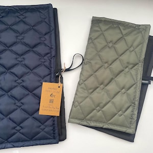 May include: A navy blue and an olive green quilted mat, each featuring a diamond-stitched pattern and a black fabric loop on the side. A rectangular brown paper tag is attached to the navy mat with the text Handmade Pocket Sit Mat.