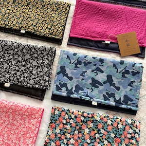 May include: A collection of rectangular fabric mats featuring various patterns including floral prints, camouflage, and polka dots. Each mat has a solid black underside and a small white label. A brown tag with text is attached to the pink mat.