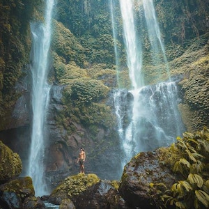May include: A scenic photograph of a waterfall cascading down a lush, green cliffside. A person stands on a moss-covered rock in the foreground, with the water flowing into a pool below. The image captures the beauty of nature.
