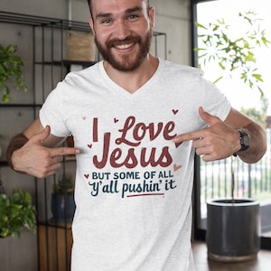 May include: White V-neck t-shirt with the text "I Love Jesus but some of all y'all pushin' it" in burgundy and teal. The shirt has small heart accents and is made of a soft material.