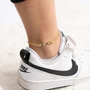 May include: A gold anklet with a heart charm and colorful gemstones. The anklet is worn on a person's ankle with white sneakers.