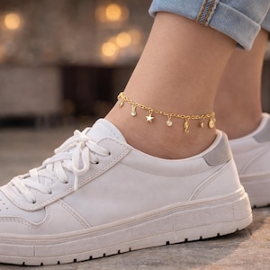 May include: A gold-colored anklet with charms of stars, circles, and seahorses. The anklet is worn on an ankle above a white sneaker with gray accents. The background is blurred with warm tones.