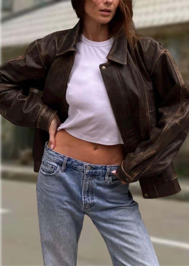 Op de afbeelding: Een bruine leren jas met een kraag en knoopsluiting. Gedragen over een wit t-shirt en gecombineerd met lichtblauwe jeans. De outfit is casual en stijlvol.