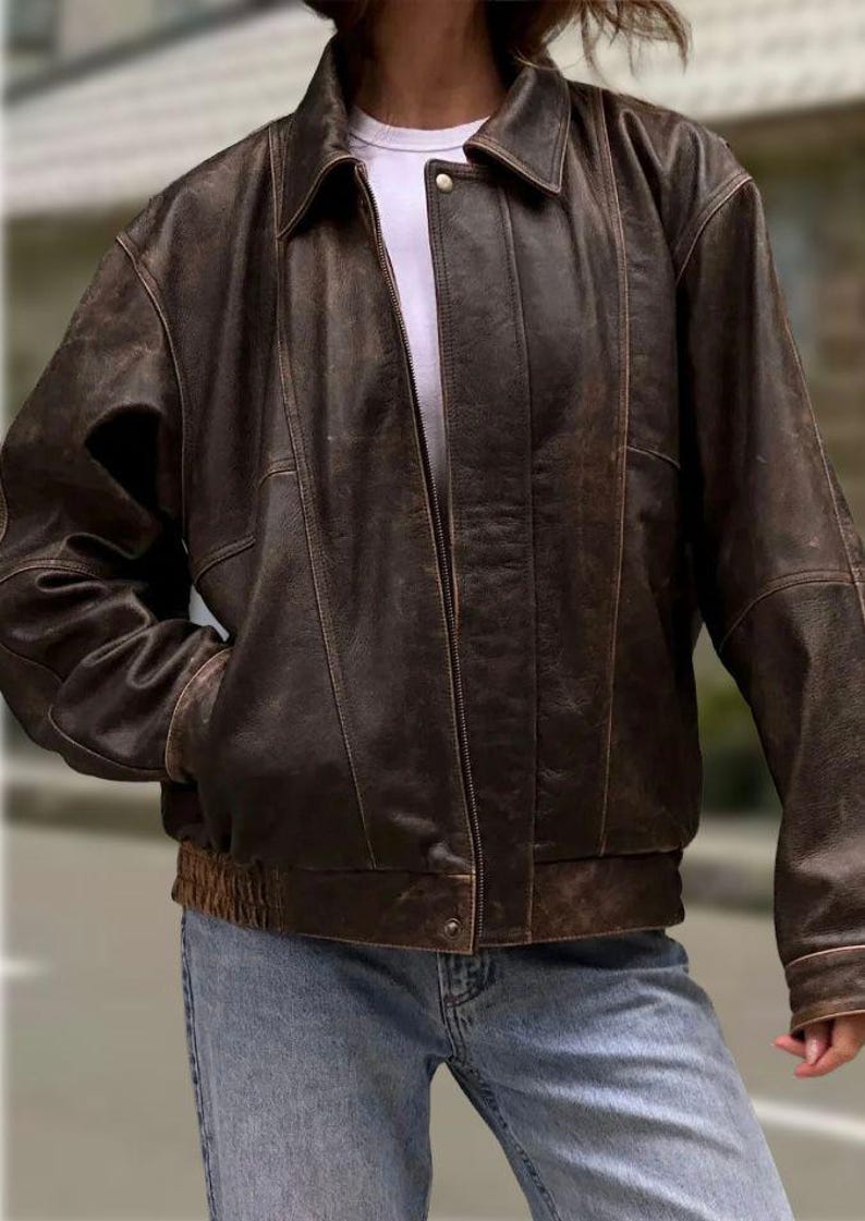 Op de afbeelding: Een bruine leren jas met ritssluiting, kraag en geribde zoom. De jas heeft een vintage, distressed look en heeft twee voorzakken. De mouwen hebben een geribde manchet. De jas wordt gedragen over een wit t-shirt en een blauwe jeans.