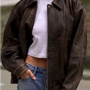 Op de afbeelding: Bruine leren jas met een distressed afwerking. De jas heeft een kraag, knoopsluiting en zakken. Gedragen over een wit t-shirt en een blauwe jeans.