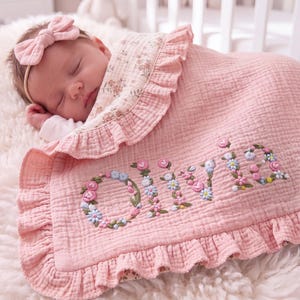 Pode incluir: Uma manta de bebé em musselina rosa suave com uma borda franzida e o nome OLIVIA bordado numa fonte floral. A manta tem uma superfície de tecido texturizado e é combinada com uma fita de cabelo com laço rosa a condizer.