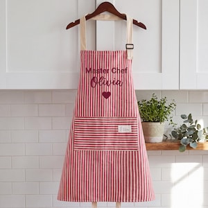 May include: A red and white vertically striped apron featuring the embroidered text Master Chef and Olivia above a small heart icon. The apron includes a large front pocket with a small rectangular label and beige neck straps.