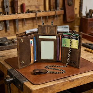 以下が含まれることがあります： 茶色の革製財布で、複数のカードスロットと透明なIDウィンドウが表示されています。財布にはチェーンが取り付けられ、「KRS」のイニシャルがエンボス加工されています。茶色の革製のマットの上に置かれています。