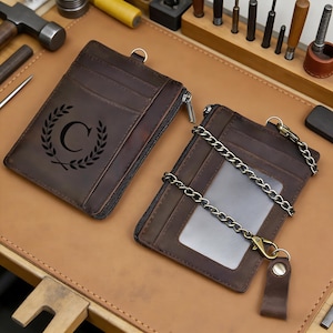 以下が含まれることがあります： 茶色の革製カードケース2つ。ジッパーと金属チェーンのディテール。1つは黒い月桂樹のデザインと文字「C」が特徴。もう1つはID用の透明な窓があります。どちらも金属リングが付いています。