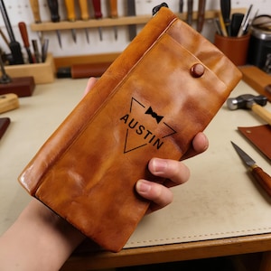 以下が含まれることがあります： 「Austin」の文字と蝶ネクタイのグラフィックが入った茶色の革製ケース。ケースはボタン留めとテクスチャ加工された表面が特徴です。ケースは手で持たれています。