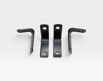 May include: Four black metal L-shaped brackets arranged in a row. Each bracket features a smooth, dark finish and two circular mounting holes, one on each perpendicular side, designed for securing joints or supporting shelves.