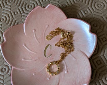 May include: A pink, flower-shaped trinket dish with a gold necklace. The necklace has a small pendant and a circular pendant with small, clear stones. The letter "C" is painted in gold on the dish.