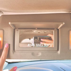 May include: A beige car visor with a built-in mirror and two small lights. The mirror displays the text "JESUS LOVES YOU" with a heart symbol. A hand with pink nail polish is touching the visor.
