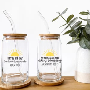 Może przedstawiać: Dwa przezroczyste szklane kubki z bambusowymi pokrywkami i słomkami. Każdy kubek ma żółtą grafikę słońca i cytat w czarnym tekście. Jeden brzmi "THIS IS THE DAY the lord has made PSALM 118:24". Drugi mówi "HIS MERCIES ARE NEW every morning LAMENTATIONS 3:22-23".