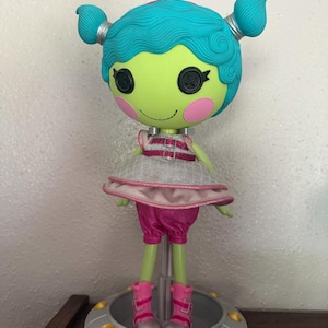 May include: A Lalaloopsy doll with a green face, teal hair, and pink accents. The doll wears a pink and silver outfit and pink boots. It stands on a silver platform with gold details, resembling a spaceship.
