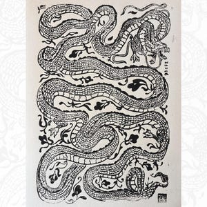 Puede incluir: Grabado en madera en blanco y negro de un dragón serpentino con escamas y cabeza detalladas. El dragón serpentea a través de un fondo de follaje estilizado. La obra de arte está sobre papel color crema.