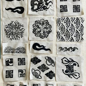 Può includere: Una collezione di piccoli campioni di tessuto quadrati con varie stampe a blocchi di inchiostro nero. I disegni includono forme astratte, motivi floreali e motivi geometrici. I campioni sono disposti su uno sfondo di tessuto chiaro.